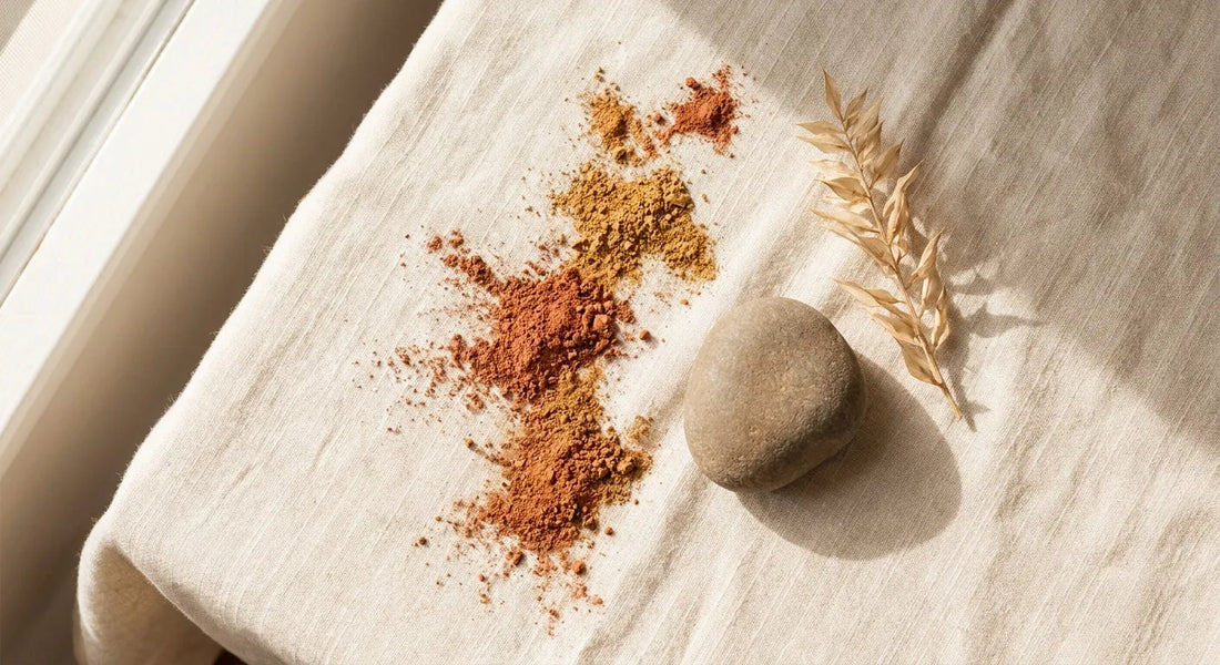 A serene flat-lay of loose terracotta and golden mineral pigment powder scattered beside a smooth stone and dried botanicals on a warm cream linen surface, lit by soft natural light.