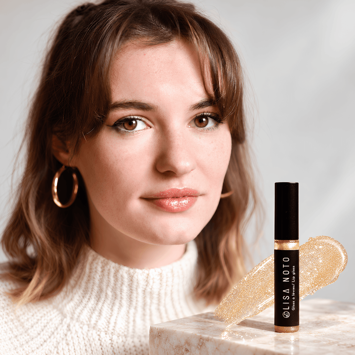 Portrait d’une femme aux lèvres rosées et brillantes, avec le gloss hydratant Lisa Noto et une touche de sa texture transparente aux paillettes dorées au premier plan.